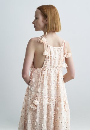 Svetloružové kvetinové šaty s viazaním na ramenách, volánikovými detailmi a voľným strihom. Majú viacvrstvový materiál s kvetinovými aplikáciami a hlboký výstrih na chrbte.