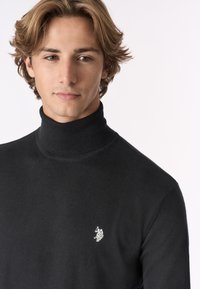 Maglione a collo alto di colore nero, realizzato in morbido tessuto a maglia, con colletto a coste e un piccolo dettaglio del logo bianco sul petto.