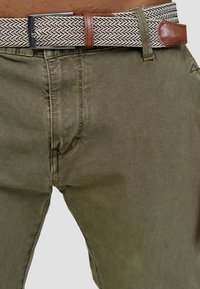 Oliivinvihreät housut, joissa on teksturoitu kudottu vyö kuvioidulla designilla ja ruskealla nahkaisella yksityiskohdalla. Kangas näyttää pehmeältä ja siinä on hillitty kiilto.