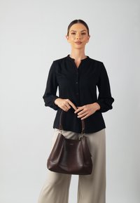 Blusa nera a maniche lunghe con chiusura a bottoni e spalle plissettate, abbinata a una borsa a secchiello in pelle marrone con forma curva e tracolla.