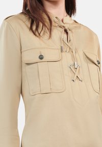 Beige long-sleeve top with front lace-up neckline and two flap chest pockets secured with decorative buttons.