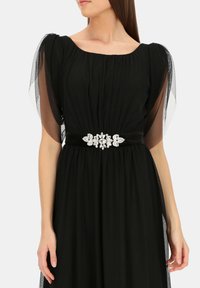 Czarna sukienka z marszczonym materiałem, przezroczystymi bufiastymi rękawami i dekoracyjnym paskiem z kryształowymi akcentami. Gładka tekstura i długość do podłogi.