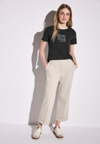 Svart t-shirt med tryckt text, beige vida byxor och vita sneakers med bruna sulor. Slät tyg, avslappnad passform och uppvikta ärmar.