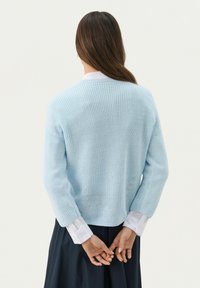 Lichtblauwe gebreide trui met een relaxte pasvorm, voorzien van lange mouwen en een ronde halslijn. Gelaagd over een witte blouse met kraag.