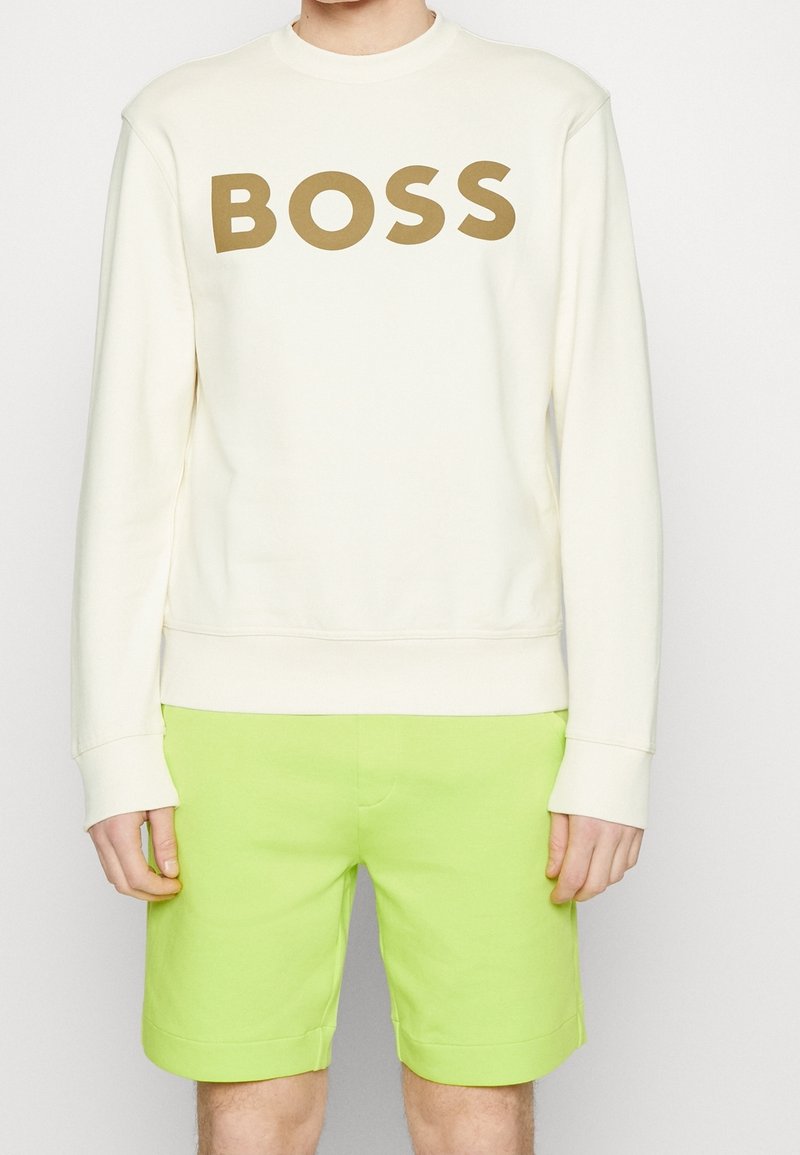 Sudadera crema con el texto "BOSS" en marrón intenso, combinada con pantalones cortos de color verde lima brillante. La sudadera tiene un cuello redondo y puños acanalados.