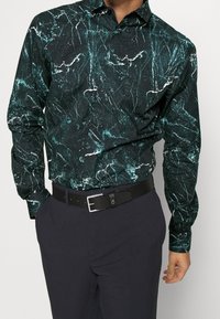 Camicia a maniche lunghe verde scuro con un motivo a marmo in turchese chiaro e bianco, abbinata a pantaloni scuri e una cintura nera.