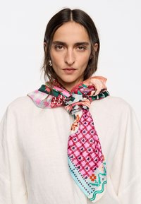 Pañuelo de seda con vibrantes patrones florales y geométricos en rosa, verde y naranja, atado en un nudo alrededor del cuello del modelo.