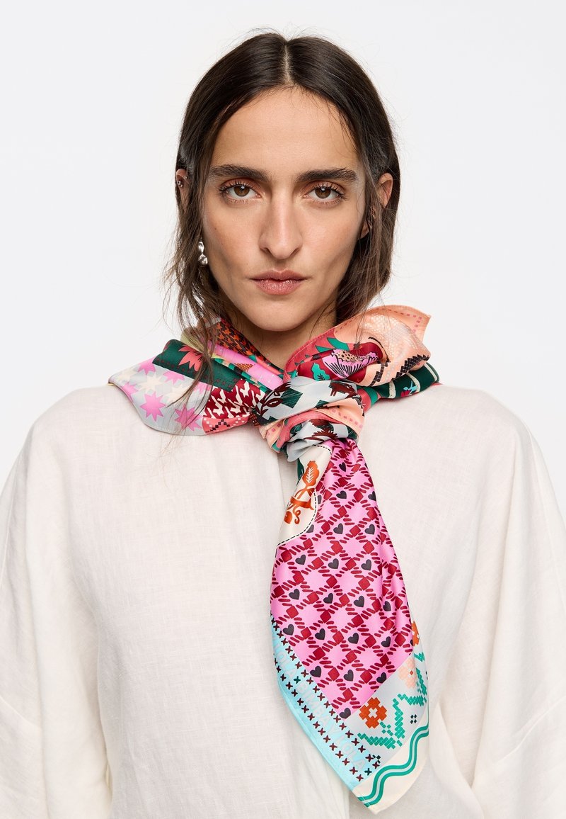 Pañuelo de seda con vibrantes patrones florales y geométricos en rosa, verde y naranja, atado en un nudo alrededor del cuello del modelo.