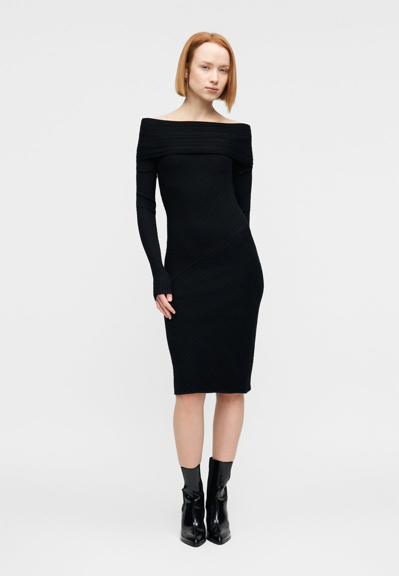 Γυναίκα με κοντά κόκκινα μαλλιά, φορώντας εφαρμοστό, μαύρο ριμπ φόρεμα χωρίς ώμους και μαύρα μποτάκια, στέκεται μπροστά σε λευκό φόντο.