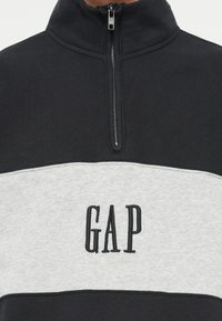 Pulóver s polovičným zipsom v čiernej farbe so sivým pruhom. Obsahuje vyšitý logotyp "GAP" na hrudi a hladkú, textúrovanú tkaninu.