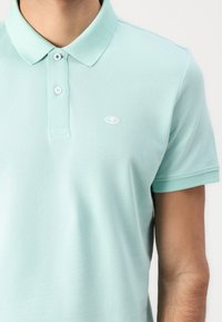 Heleheles roheline polo-särk, mis on valmistatud tekstureeritud puuvillast, klassikalise kraega, kahe nupuga kitsas ja väikese tikandiga logo rinnal.