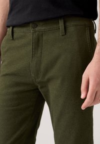 Person wearing dark olive green wool trousers with button and belt loops, paired with a black shirt, hand resting by side.