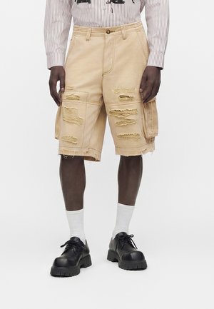Beige cargo shorts ned til knæet med flere slidte lapper, båret med sorte sko, hvide sokker og en langærmet, stribet skjorte.