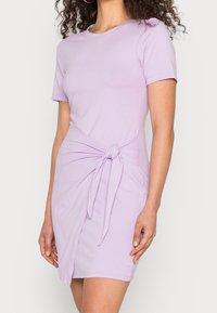 Lila kortärmad klänning med en figurnära silhuett, som har en knutdetalj i midjan och mjuk bomullstyg.