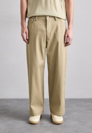 Beige cotton trousers with a relaxed fit, featuring a zip fly, front pockets, and a smooth texture. Worn with cream shoes.