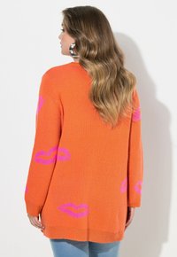 Strickstrickjacke in leuchtendem Orange mit übergroßem pinkfarbenem Lippenmuster. Mit langen Ärmeln, entspanntem Sitz und abgerundetem Saum.