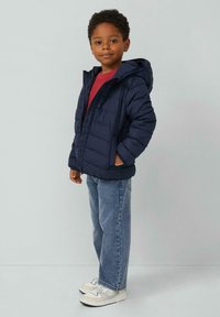 Navy pufferjack met een capuchon, met een ritssluiting en zijzakken. Gecombineerd met een rood shirt en lichtblauwe jeans, en witte sneakers.