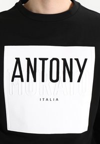 Camisa negra de manga larga con un gráfico rectangular blanco que presenta un texto en negrita negro "ANTONY" y "MORATO ITALIA" debajo. Acentos texturizados.