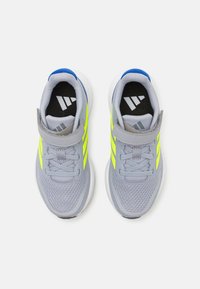 Baskets de sport grises avec une tige en maille respirante, des accents jaune fluo, un talon bleu et une sangle à crochets et boucles. Présente une semelle rembourrée.