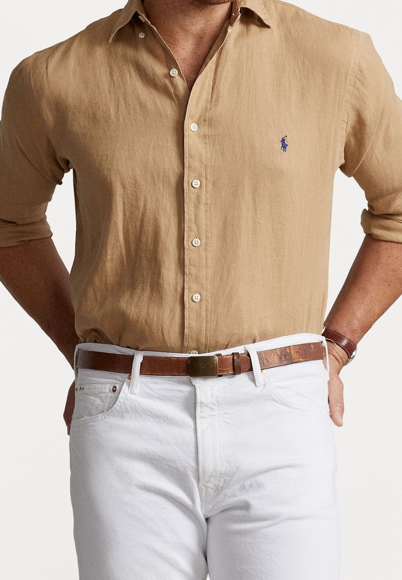 Brun knäppskjorta med uppkavlade ärmar, vita denimbyxor och ett brunt läderbälte. Har en liten blå logotyp på skjortan.