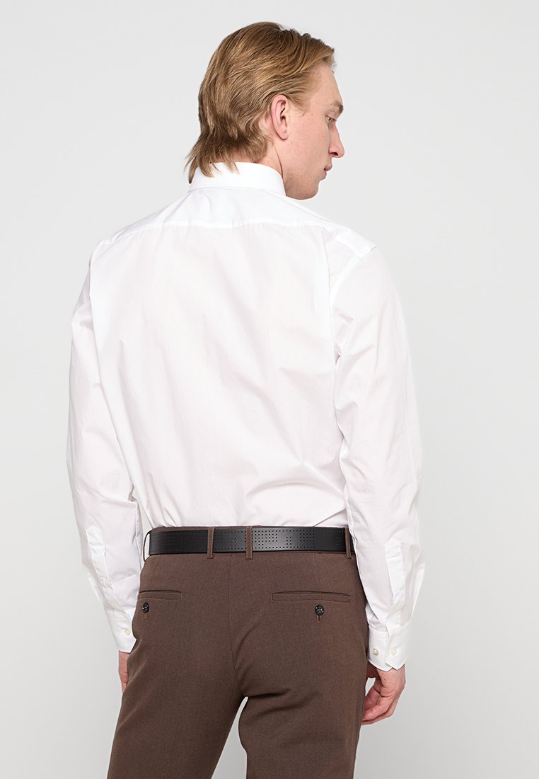 Homme portant une chemise blanche à manches longues rentrée dans un pantalon marron avec une ceinture noire, légèrement tourné sur le côté contre un fond uni.