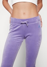 Lila velourbyxor med dragsko i midjan och metallspetsar. De har en liten broderad logotyp på framsidan. Slät textur.