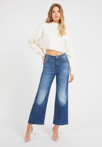 Pull blanc court avec texture côtelée, associé à un jean bleu taille haute à jambes larges présentant un délavage et un ourlet effiloché, complété par des talons transparents.
