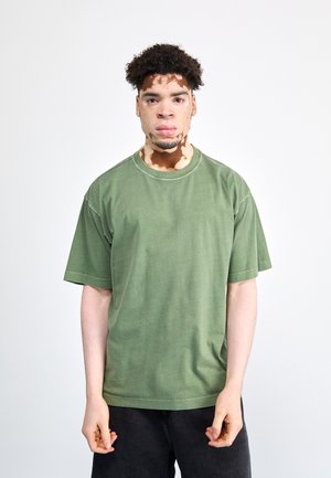 Camiseta verde caqui de gran tamaño con cuello redondo, mangas cortas y textura suave. Combinada con jeans oscuros sobre un fondo liso.