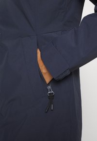 Main partiellement insérée dans la poche zippée d'une veste bleu marine avec poignet ajustable et cordons de serrage.