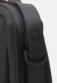 Manico di borsa sintetica nera con finitura testurizzata, caratterizzato da dettagli cuciti e un emblema circolare nero con un design a raggi.