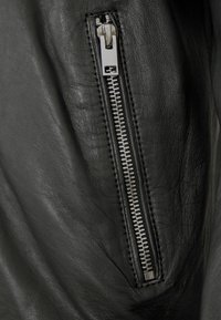 Detail einer schwarzen Lederjacke mit einem silbernen Metallreißverschluss, strukturiertem Leder und sauberer Naht entlang der Kante.