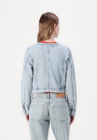 Τζάκετ από ελαφρύ τζην με cropped σχέδιο, που διαθέτει πορτοκαλί ραφές, μακριά μανίκια και fitted cuffs. Η πίσω πλευρά δείχνει έναν απλό λαιμό.