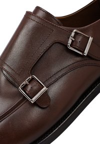 Bruine leren schoenen met een gladde textuur, voorzien van twee zilveren gespen en decoratieve stiksels. Platte zool met een rond toelopend design.