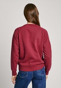 Sweatshirt bordeaux con maniche raglan e polsini a coste, abbinato a jeans di denim blu. Il tessuto sembra morbido con una superficie liscia.