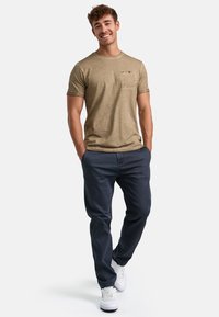 Olivengrøn tekstureret t-shirt med brystlomme, parret med mørkeblå bukser og hvide sneakers. Modellen står med hænderne i lommerne.