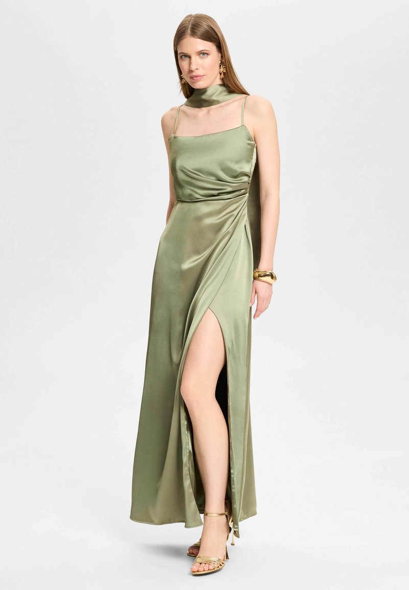 Femme portant une robe sans manches en satin vert olive avec une fente haute, bracelet doré, boucles d'oreilles et talons à brides, debout devant un fond uni.