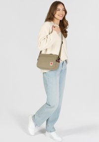 Borsa a tracolla beige con chiusura a zip, realizzata in materiale tessile, con una piccola etichetta con logo. Il modello indossa un maglione crema e jeans azzurri chiari.