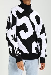 Schwarz-weißer Pullover mit hohem Kragen, kräftigen geometrischen Mustern und drei Streifen an den Ärmeln. Weicher Stoff mit lockerem Schnitt.