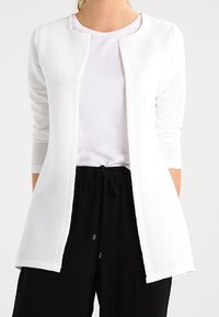 Weißer langärmeliger Cardigan mit strukturiertem Design, offener Front und lockerem Sitz, kombiniert mit einem basic T-Shirt und schwarzen Hosen.