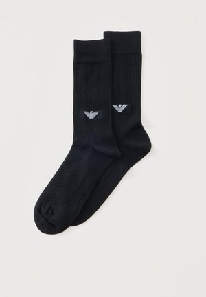 Paire de chaussettes noires côtelées montantes avec un petit logo stylisé d'aigle blanc près de la cheville sur un fond clair uni.