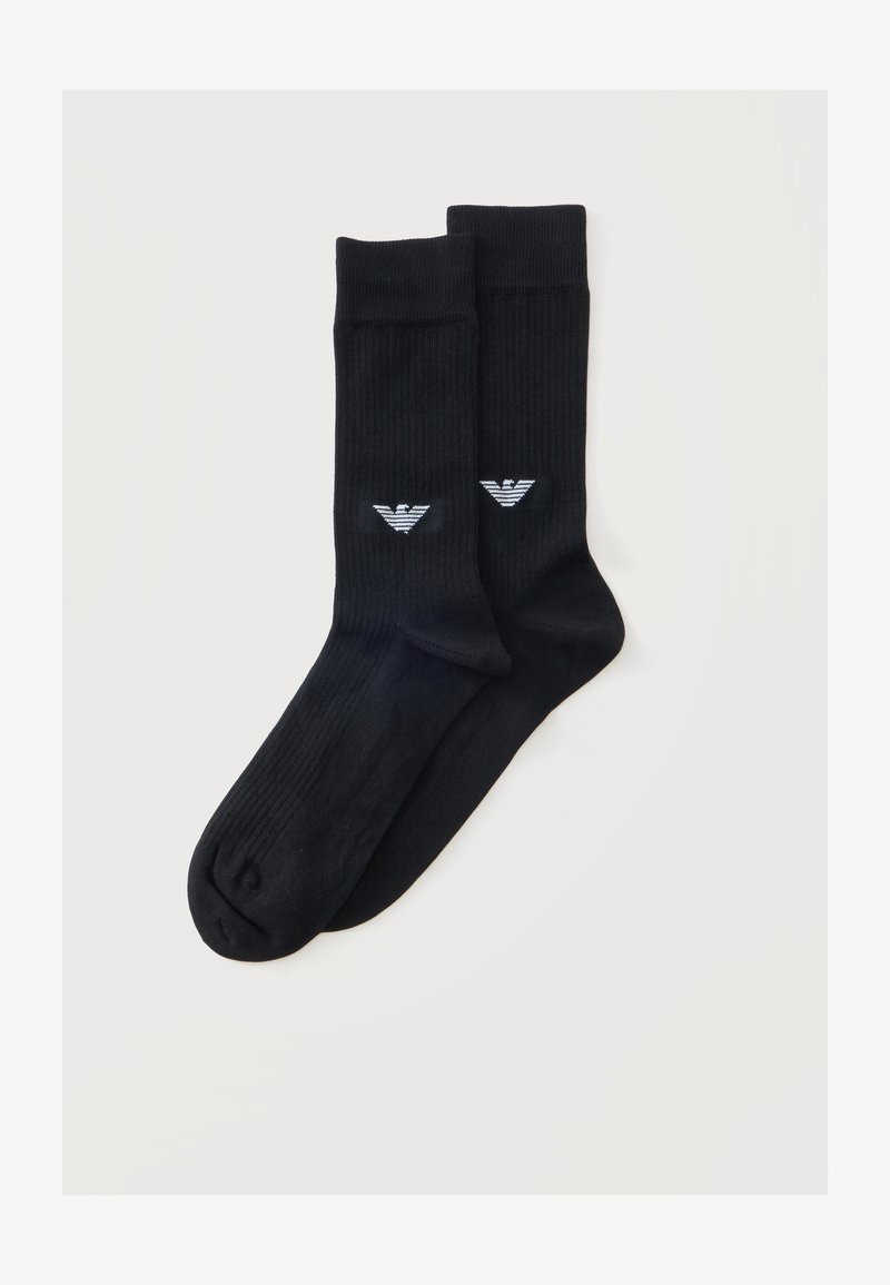 Paire de chaussettes noires côtelées montantes avec un petit logo stylisé d'aigle blanc près de la cheville sur un fond clair uni.