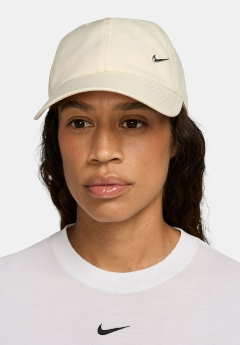 Gorra de béisbol Nike beige con visera curva, tela suave y pequeño logotipo negro en la parte frontal. Se lleva con una camiseta blanca que presenta un logotipo similar.