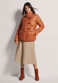 Pufferjack in verbrande sinaasappel met een hoge kraag, voorzien van zwarte toggle-sluitingen en een doorgestikt ontwerp. Gecombineerd met een beige midi-jurk en roodbruine laarzen.