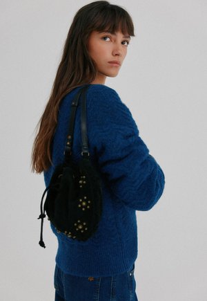 Black drawstring bag with textured surface and gold metal accents, worn over a blue knitted sweater. The bag features adjustable straps.
