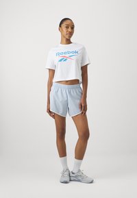 Reebok baltas trumpas marškinėliai su mėlynu logotipu, derinami su šviesiai mėlynomis sportinėmis šortais, kuriuose yra elastinga juosmuo, ir pilkais sportbačiais.