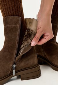 Mano desabrochando una bota de ante marrón con forro de piel, usada con gruesos calcetines de punto marrones sobre un fondo blanco.