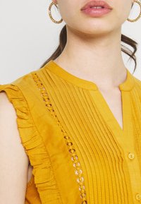 Hořčicově žlutá blůza s vertikálními sklady, volány na rukávech a dekorativním háčkováním po straně. Obsahuje knoflíkovou légou.
