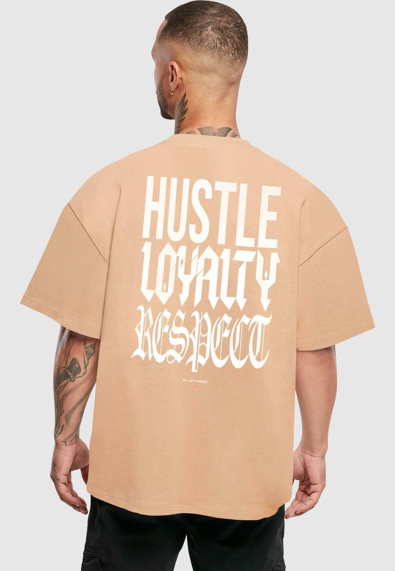 Šviesiai tan spalvos, didelių rankovių oversized marškinėliai iš medvilnės, su ryškiais baltais užrašais "HUSTLE LOYALTY RESPECT" ant nugaros.
