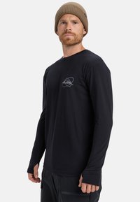 Zwarte longsleeve shirt met een ronde hals en een grijs logo op de borst. Zachte stof met een slim fit. Gepaard met donkere broek.