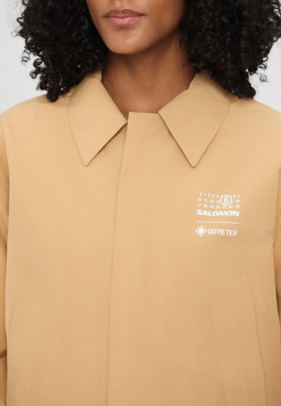 Beige Gore-Tex jakke med en stor krave og et hvidt logo med kalendertryk på brystet. Glat tekstur med minimale syningsdetaljer.